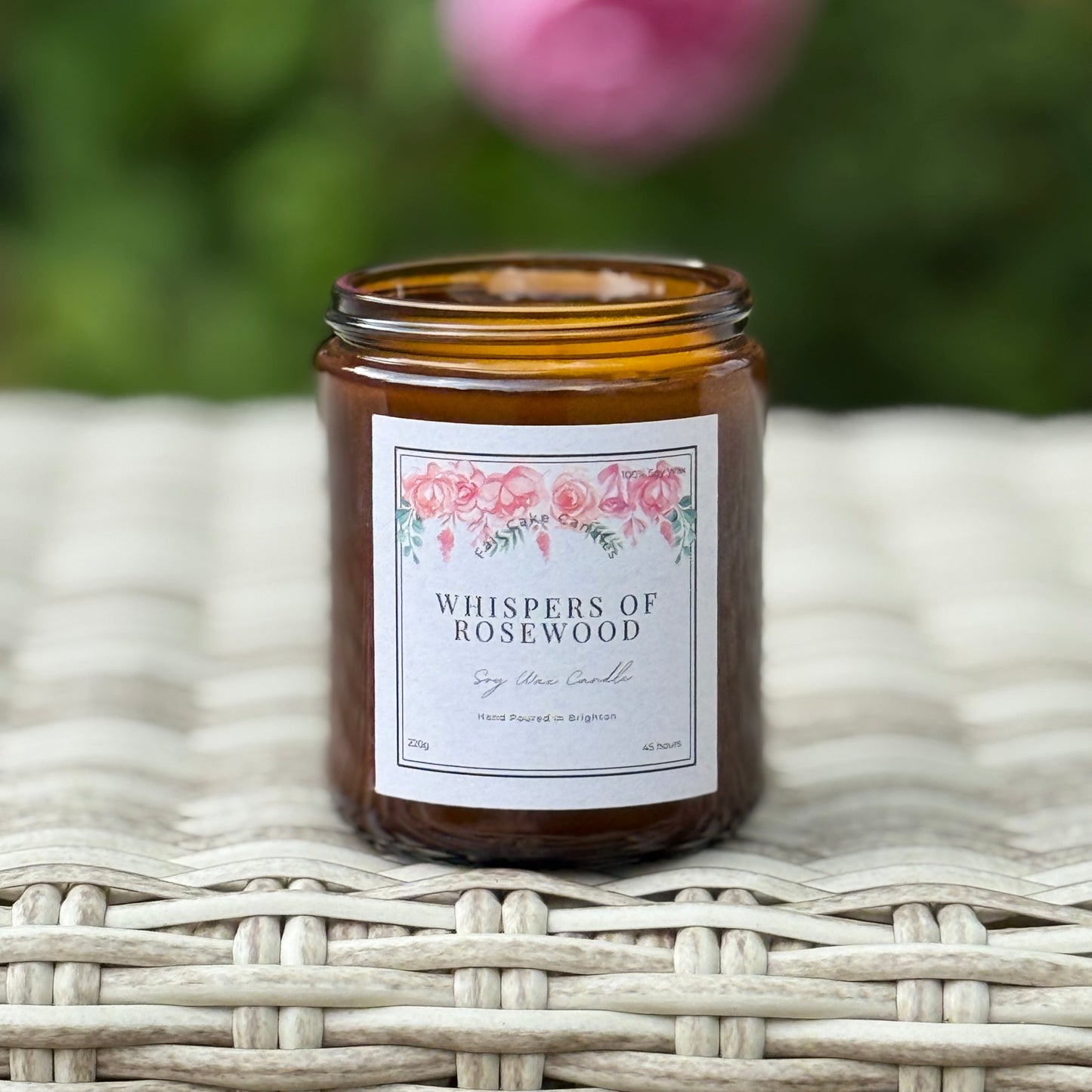 Candle jar labeled 'Whispers of Rosewood' on a woven surface with a blurred floral background