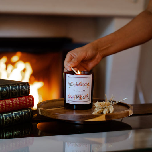 Hand lighting a 'Hello Fall' candle on a wooden tray with a fireplace in the background
