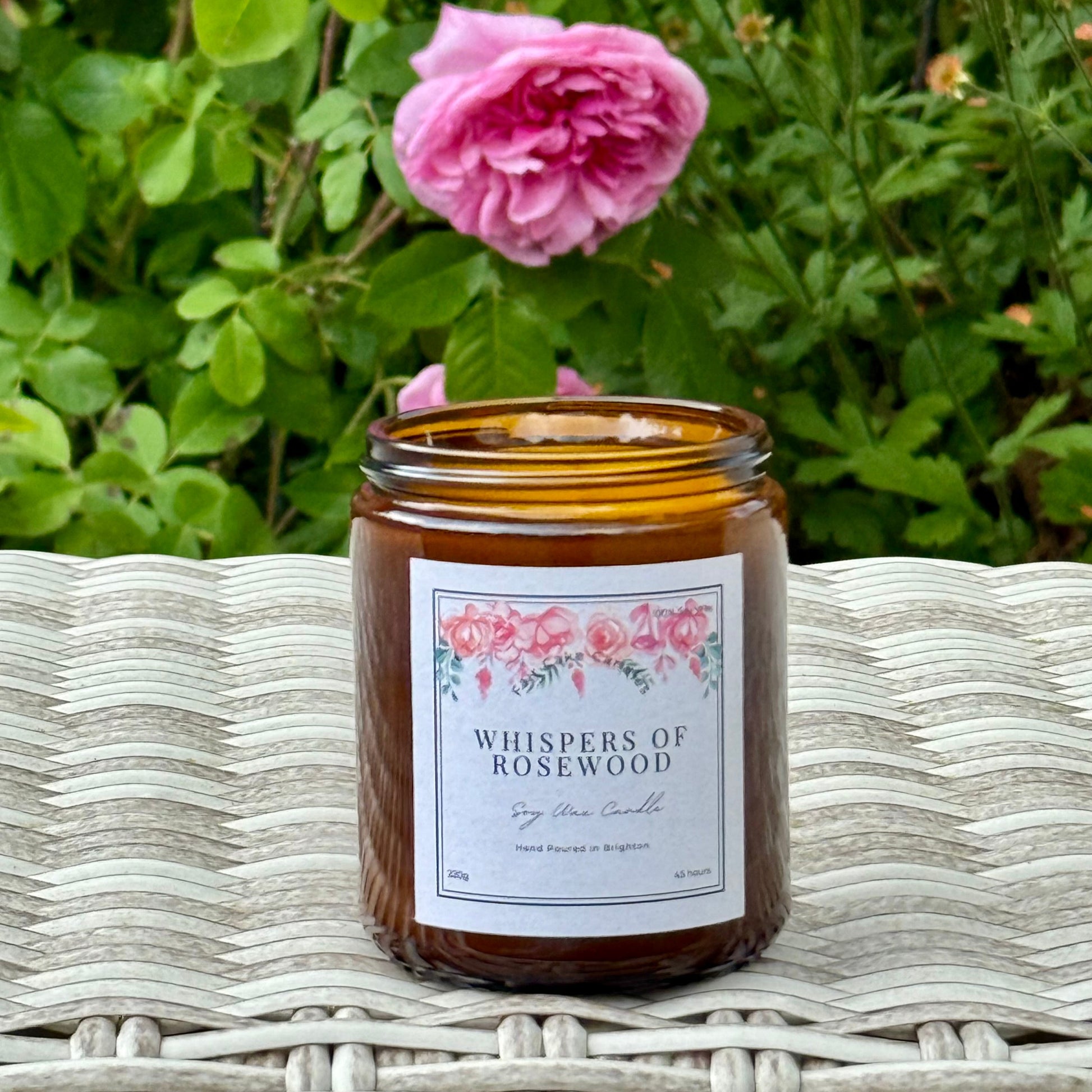 Candle labeled 'Whispers of Rosewood' on a wicker surface with pink flowers in the background