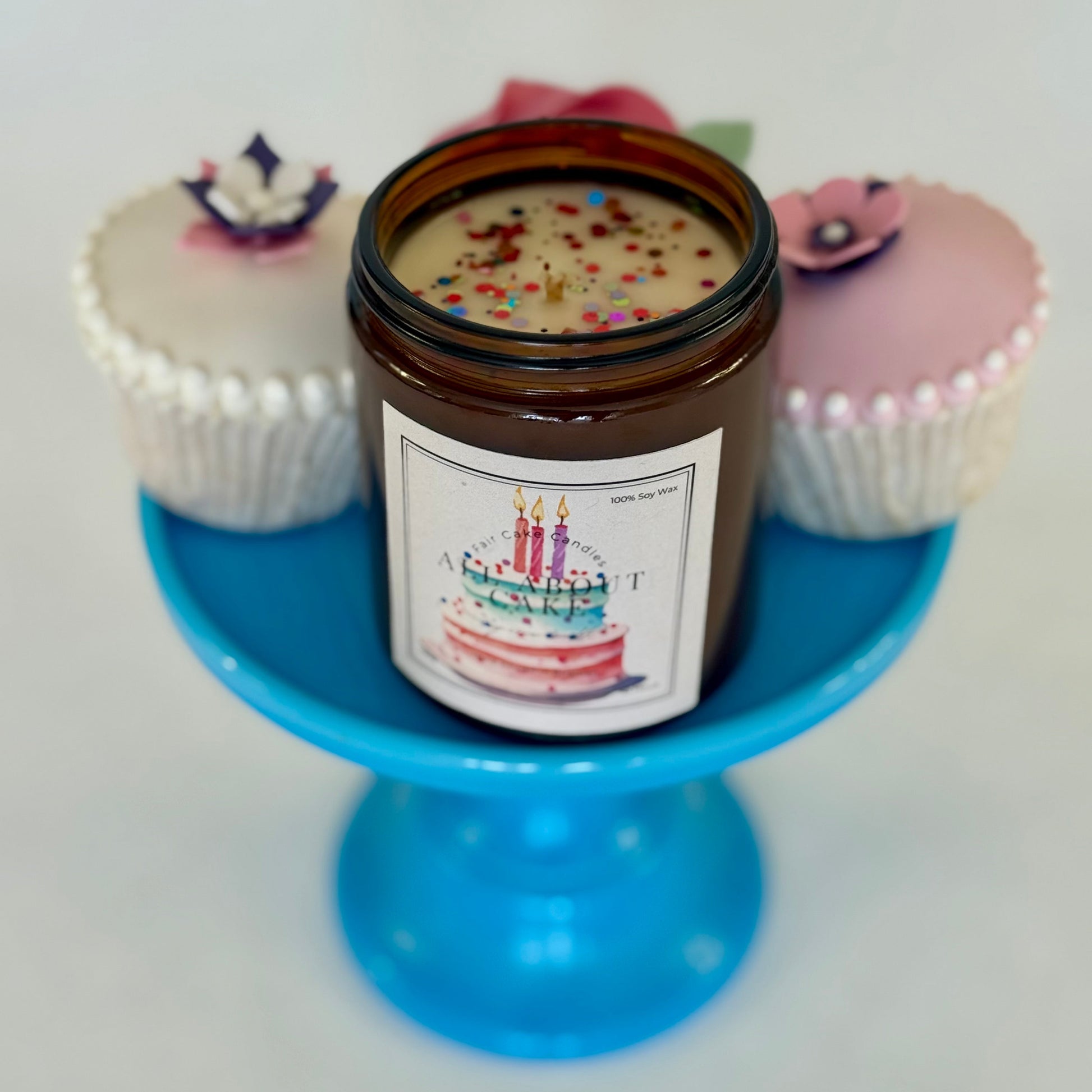 All About Cake candle with a label featuring birthday cake and candles on a blue stand with cupcake decorations.