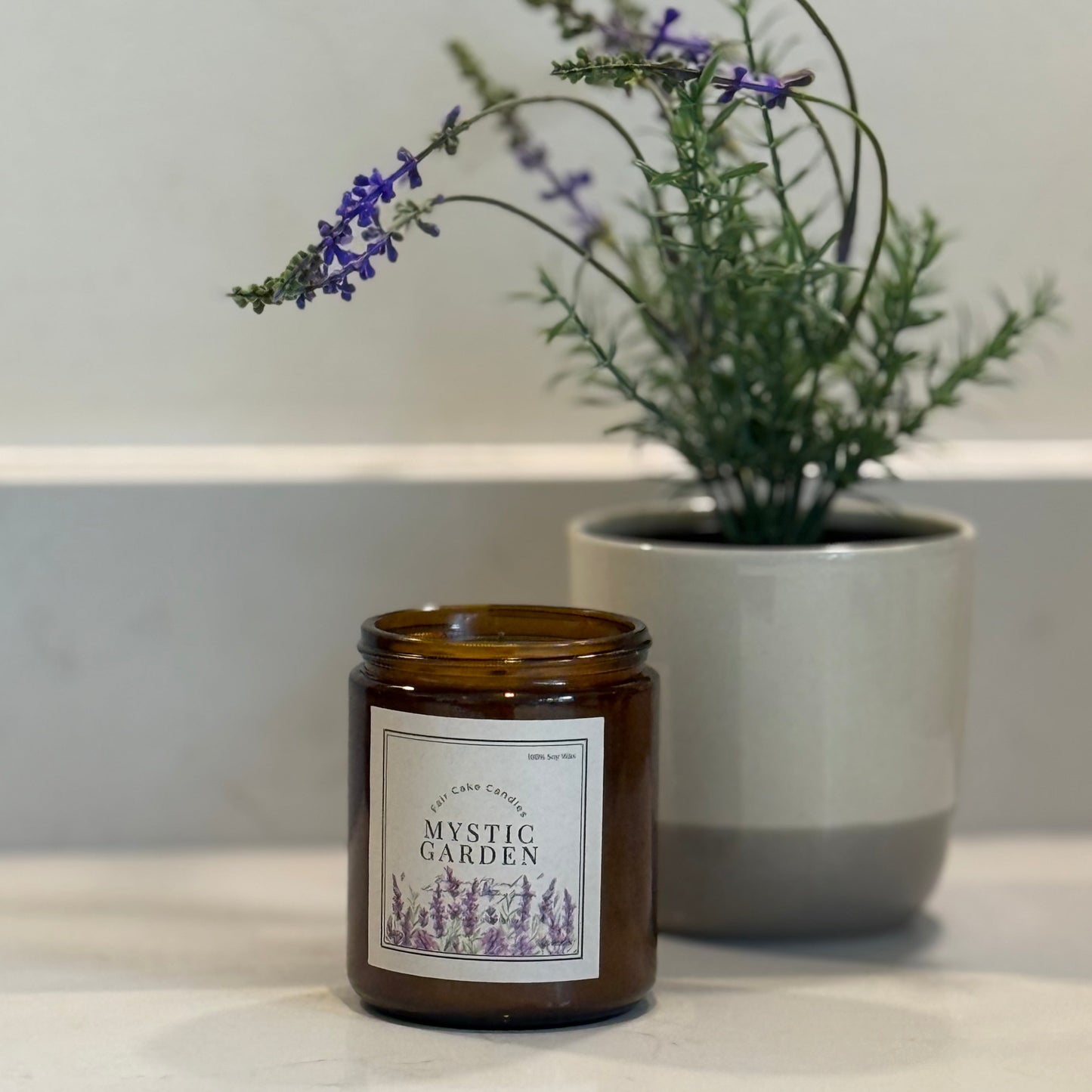 Candle labeled 'Mystic Garden' with a lavender plant in the background on a neutral surface