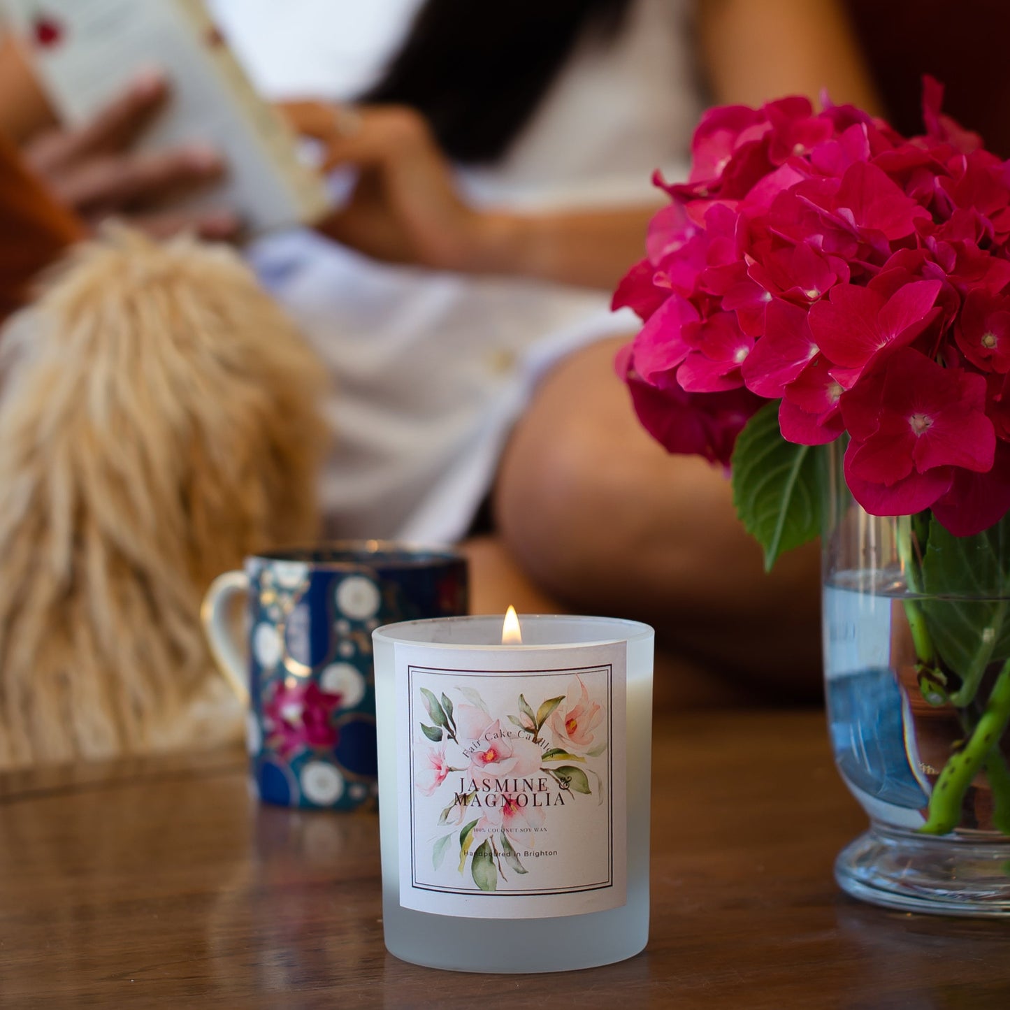 Jasmine and Magnoli botanical scented candle with a floral label on a table next to a mug and vase of pink flowers, with people in the background.