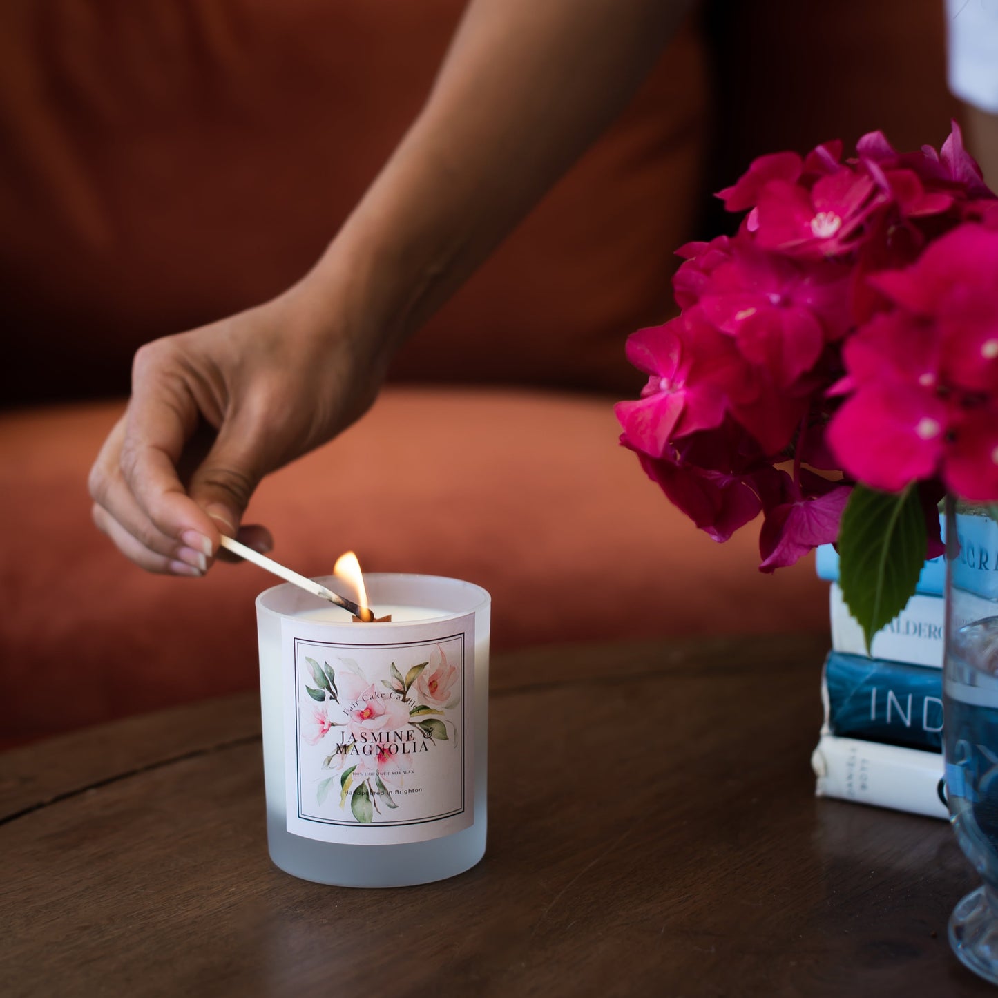 Person lighting a candle labeled 'Jasmine & Magnolia' with flowers and books in the background.