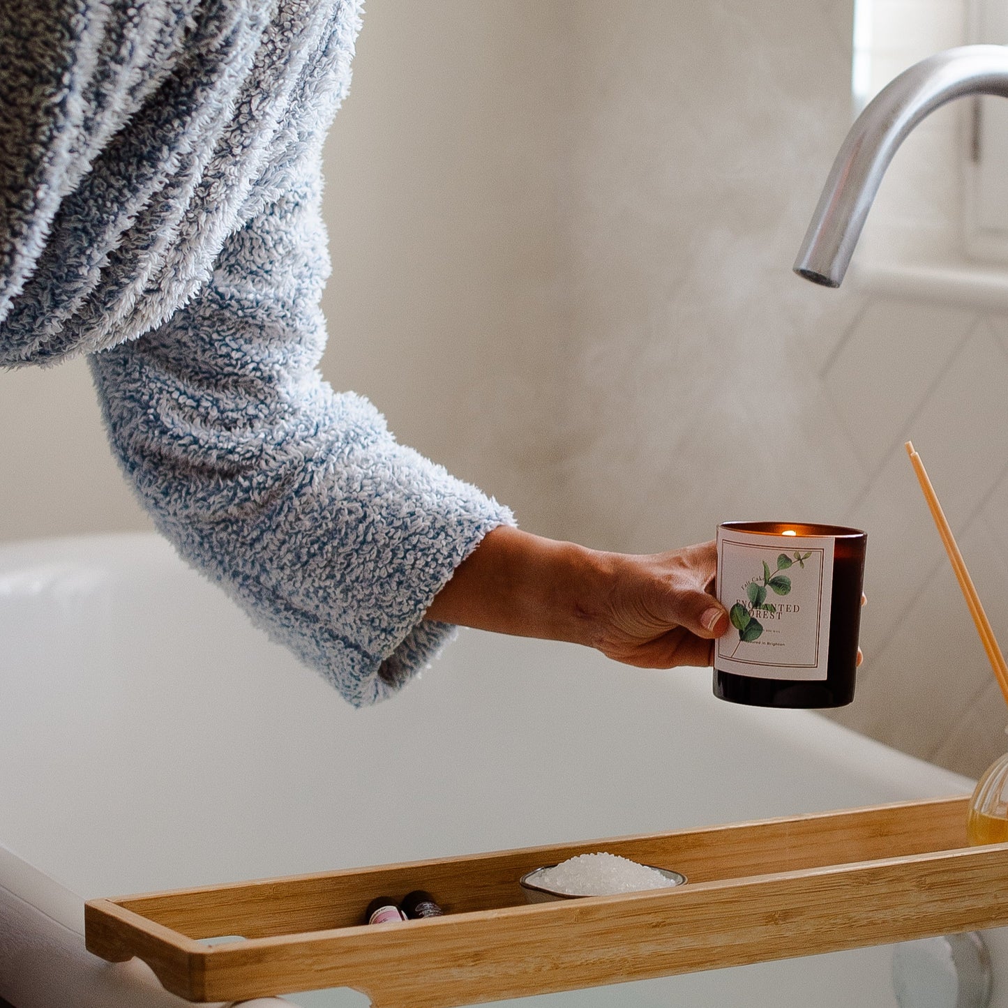 Person holding a candle over a bathtub with a wooden tray and bath items.