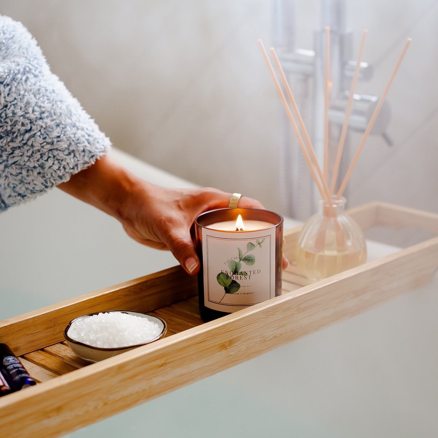 Hand holding a lit candle on a wooden tray with bath salts and a diffuser in a bathroom setting. Enchanted Forest Eucalyptus Scented Botanical Candle by Fair Cake Candles.