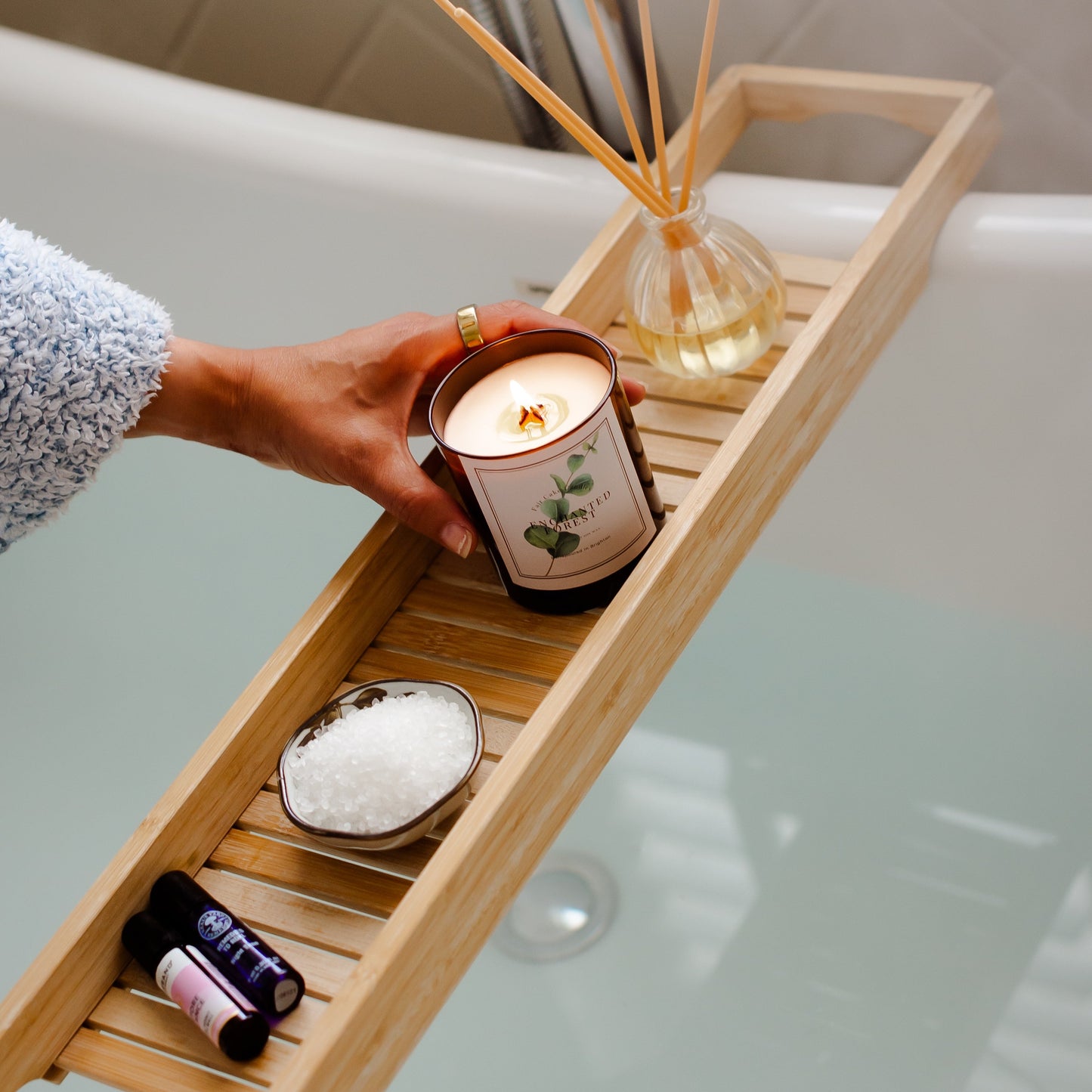 Hand holding a lit candle on a wooden bath tray with various items over a bathtub. Enchanted Forest Eucalyptus Scented Candle by Fair Cake Candles.