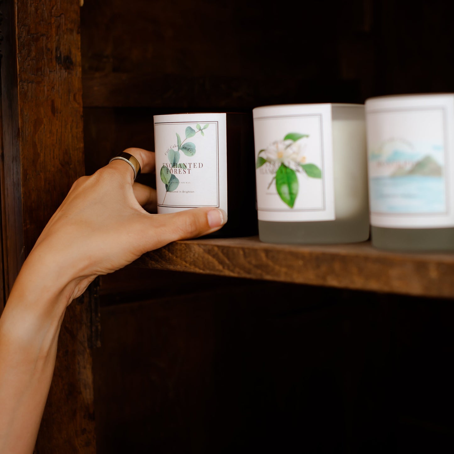 Hand holding an Enchanted Forest candle by Fair Cake Candles on a shelf with other range of candles.