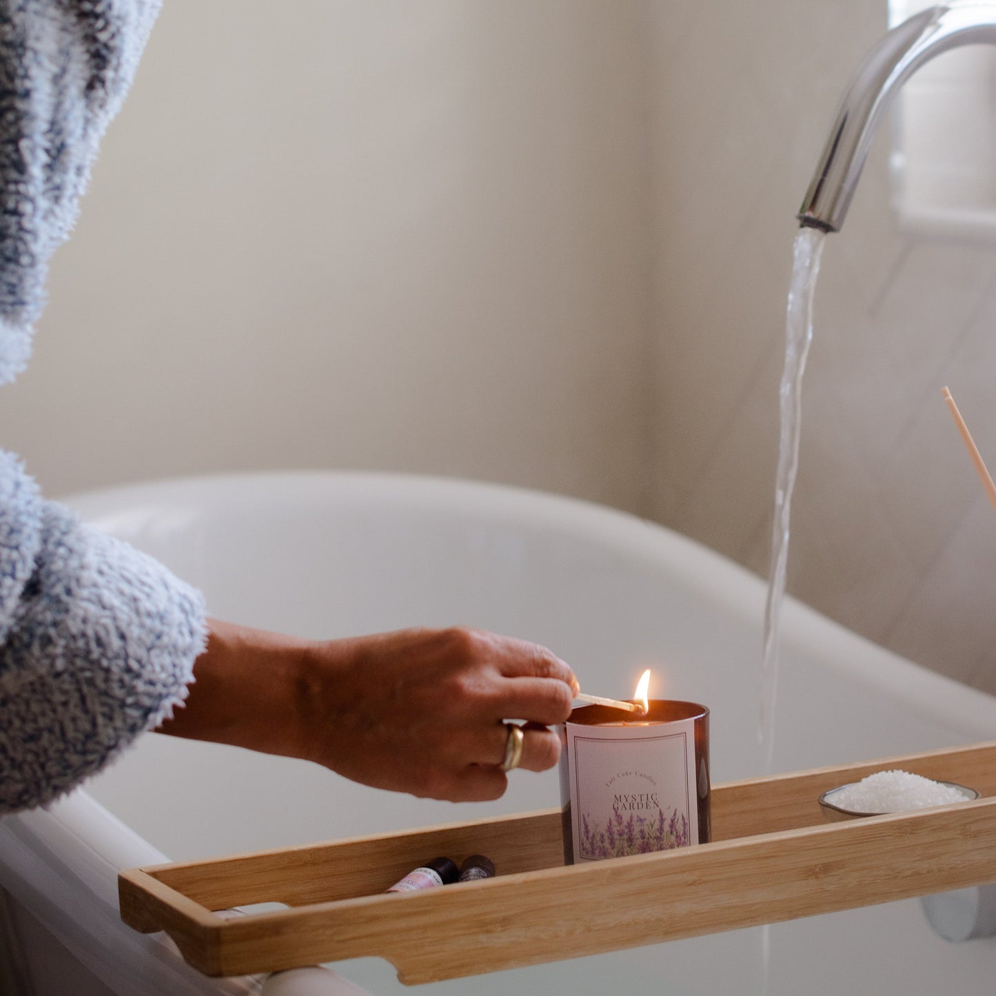 Person lighting a candle in a bathtub with a wooden tray holding a Mystic Garden Botanical Candle by Fair Cake Candles.