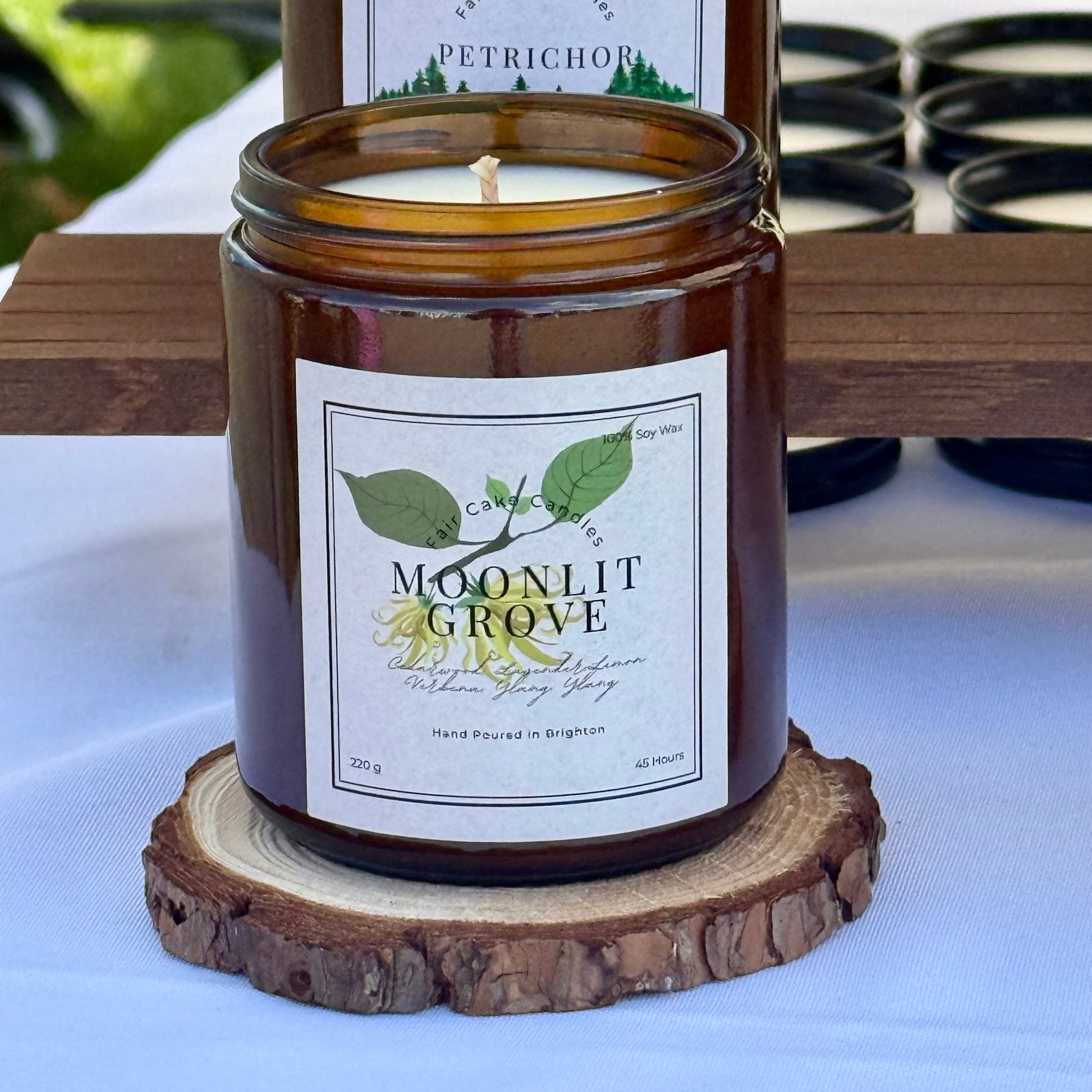 Candle in an amber jar labeled 'Moonlit Grove' on a wooden coaster with a blurred background.