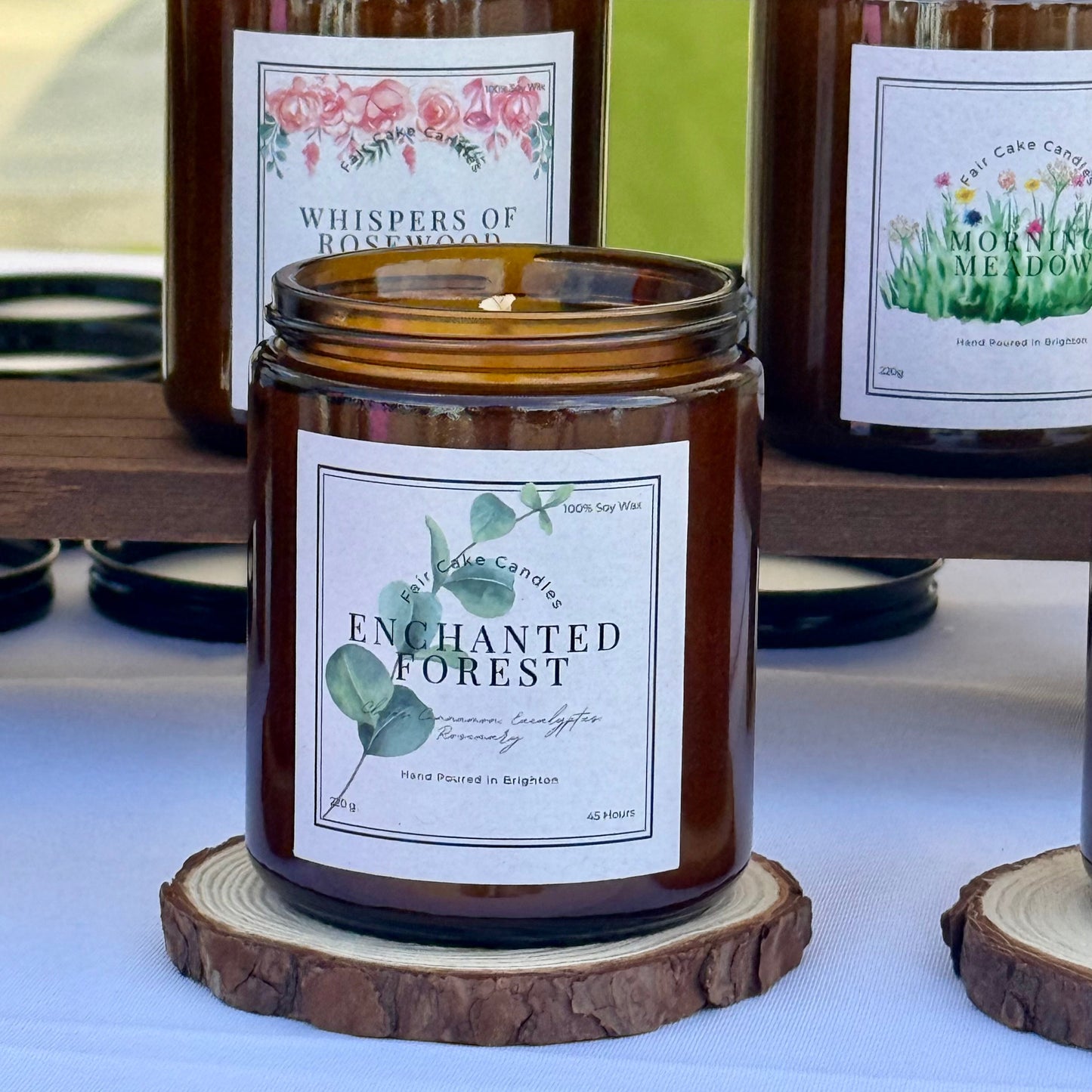 Candle jar labeled 'Enchanted Forest' on a wooden coaster with other candles in the background.