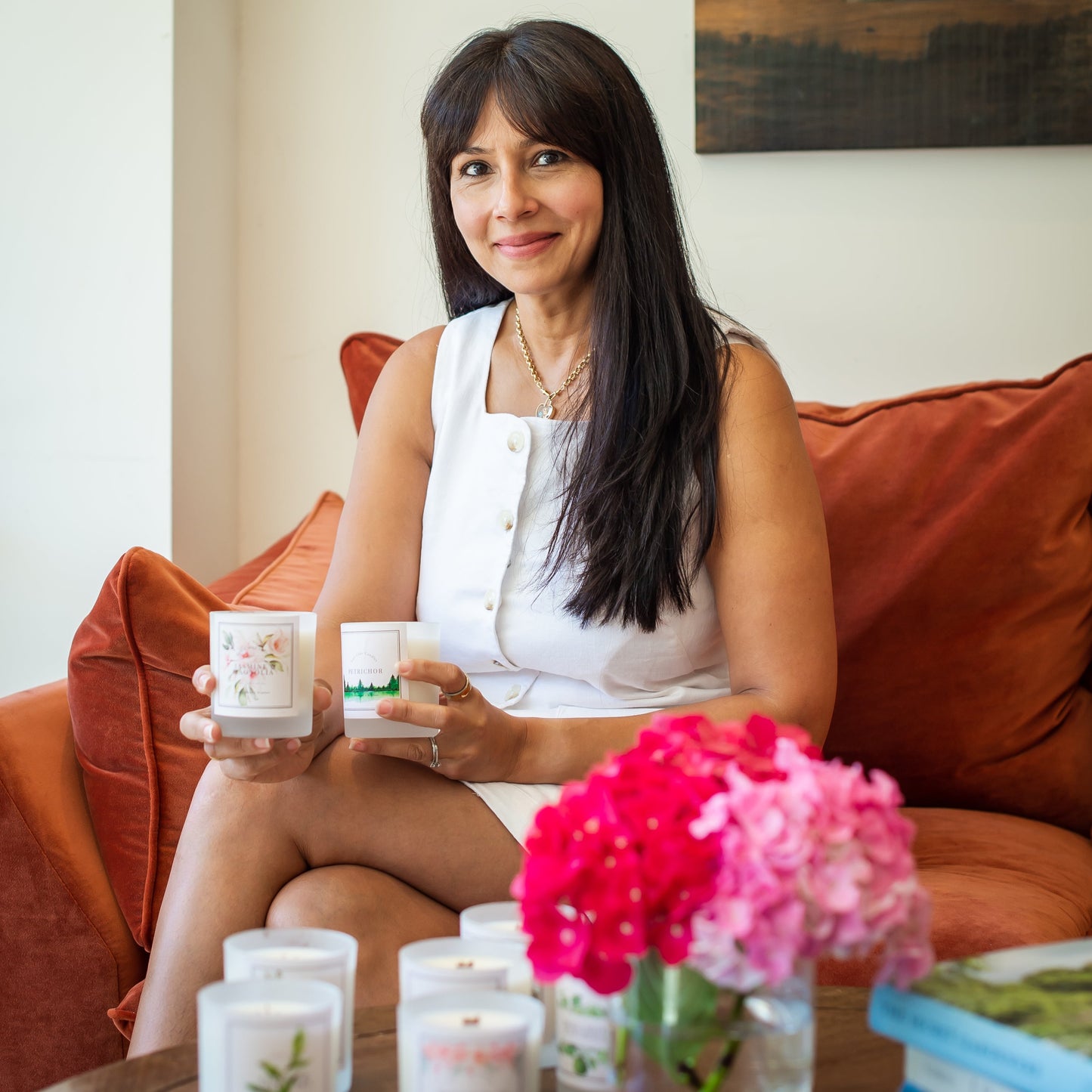 Woman sitting on a sofa holding candles with a vase of flowers in front of her.