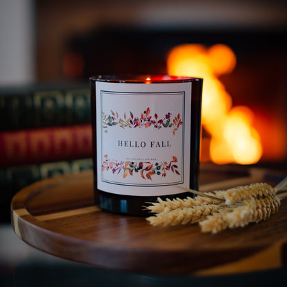 Hello Fall Candle in a glass jar on a wooden tray with blurred fireplace in the background