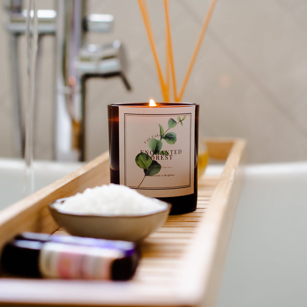 Candle labeled 'Enchanted Forest' on a bath tray with soap and books in a bathroom setting